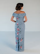 A woman shown from the back wearing a floor-length off-the-shoulder gown covered in light blue sequins with pink floral embroidery, photographed against a plain light gray studio background. / Off the shoulder column gown with all over beading and multi color detailing.