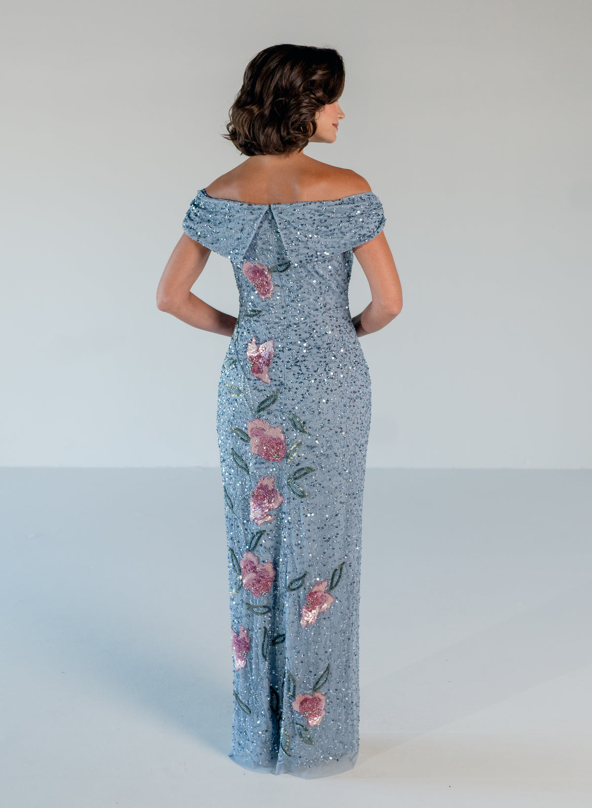 A woman shown from the back wearing a floor-length off-the-shoulder gown covered in light blue sequins with pink floral embroidery, photographed against a plain light gray studio background. / Off the shoulder column gown with all over beading and multi color detailing.
