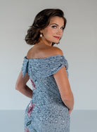 A woman wearing a light blue sequined off-the-shoulder evening gown, photographed from the back-three-quarter angle against a plain light gray studio background. / Off the shoulder column gown with all over beading and multi color detailing.