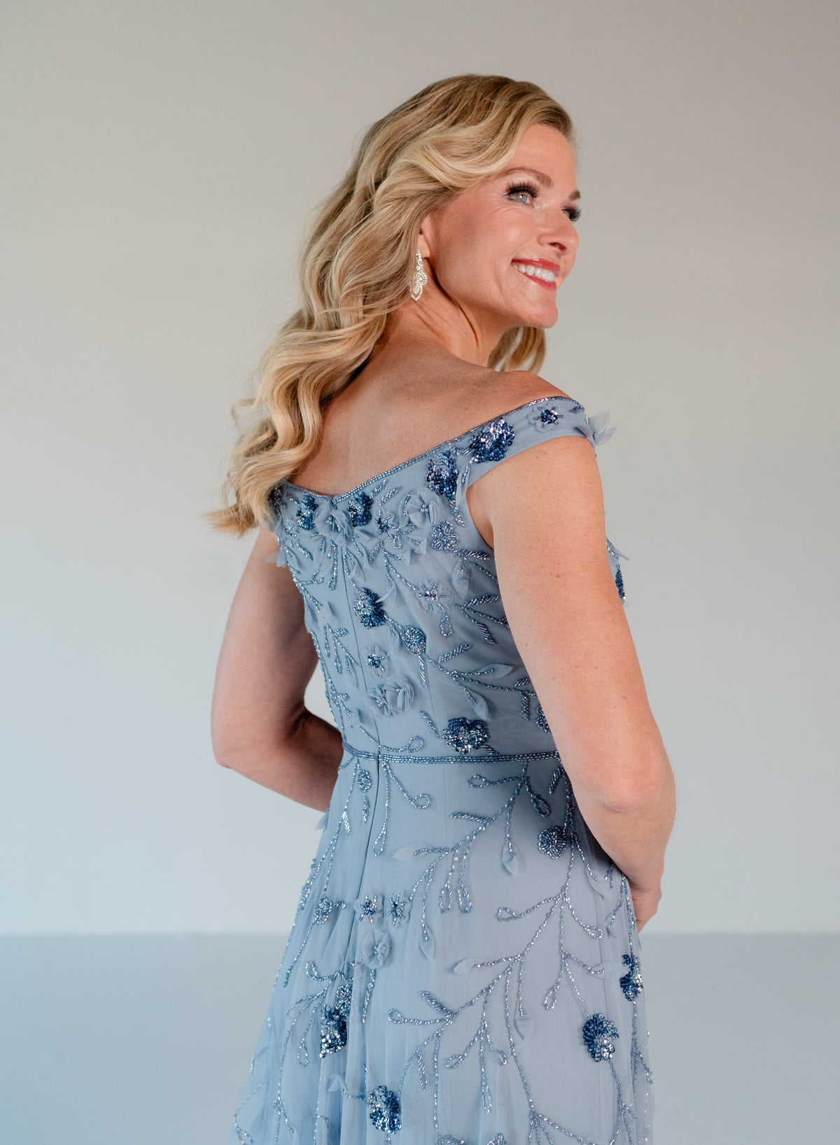 A woman wearing a light blue off-the-shoulder evening gown with intricate beaded floral details, photographed from the back against a plain light gray studio background. / Tip of the shoulder, soft A-line gown with 3D floral beading.