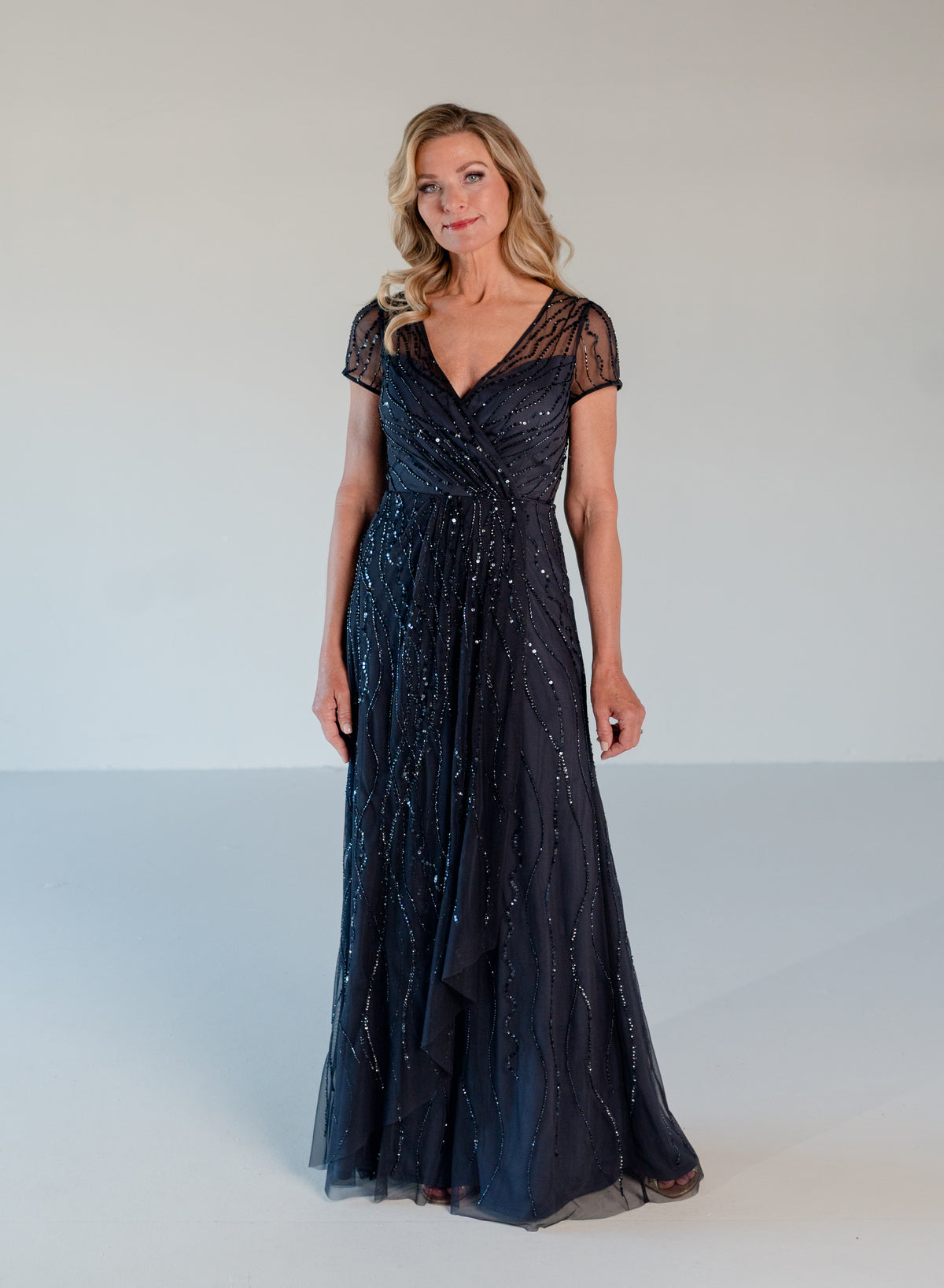 A woman modeling a floor-length navy blue evening gown with sheer short sleeves, a V-neckline, and shimmering sequin embellishments, set against a plain light gray studio background. / V neckline, A-line skirt with soft cascade and all over beading.