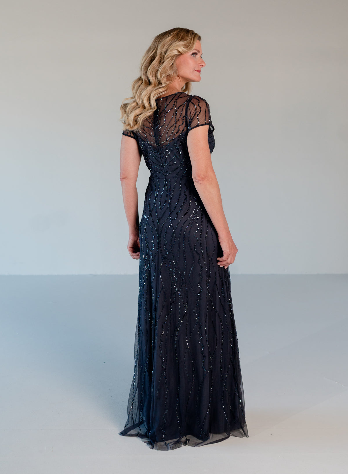 A woman modeling a floor-length navy evening gown from the back, featuring sheer cap sleeves, a lace-embroidered back, and shimmering sequin embellishments, set against a plain light gray studio background. / V neckline, A-line skirt with soft cascade and all over beading.