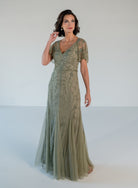 A woman modeling a floor-length sage green evening gown with short sheer sleeves, a V-neckline, and delicate floral embroidery, set against a plain white studio background. / V neckline, soft flutter sleeve gown with godet skirt and all over beading.