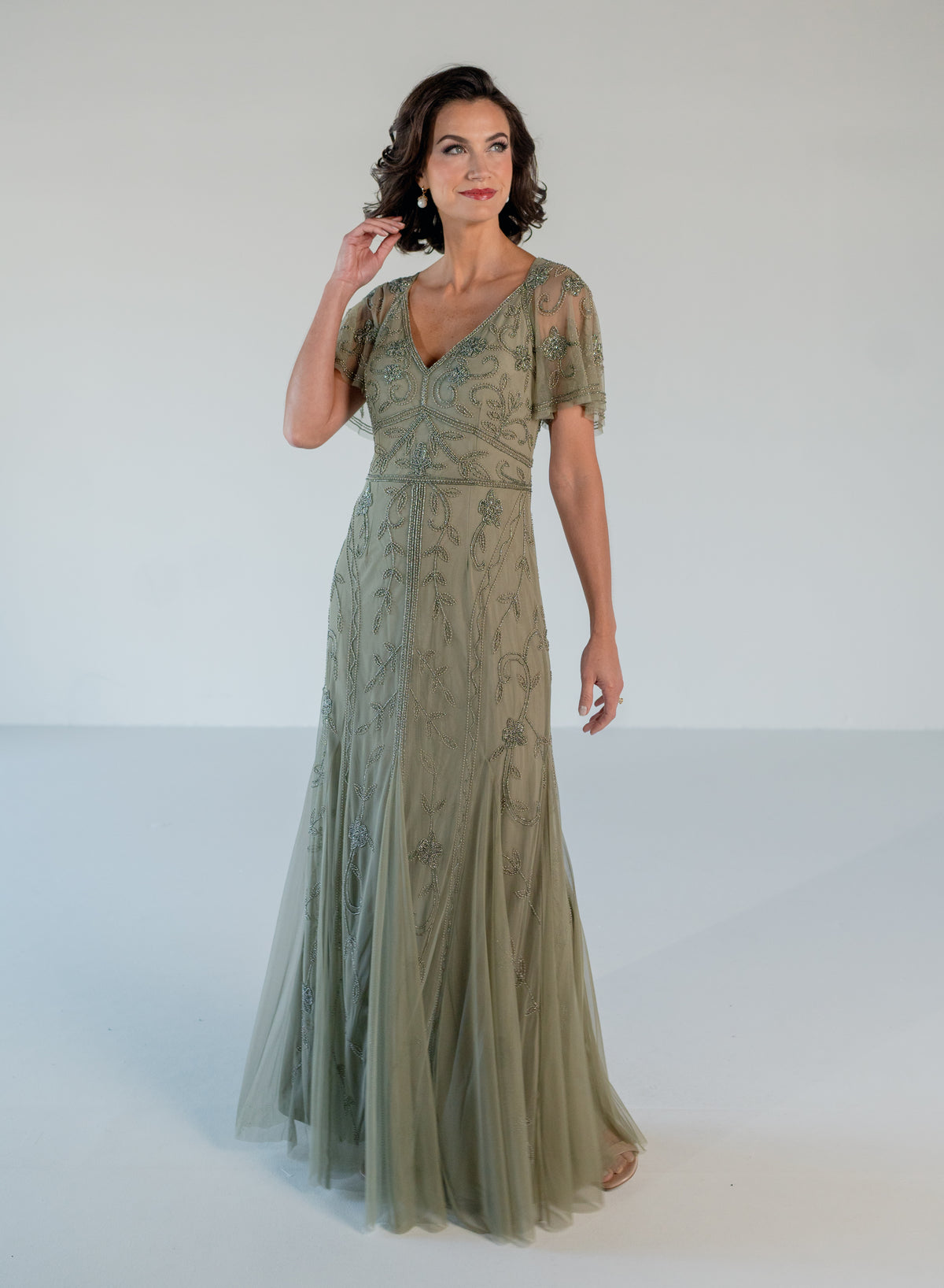 A woman modeling a floor-length sage green evening gown with short sheer sleeves, a V-neckline, and delicate floral embroidery, set against a plain white studio background. / V neckline, soft flutter sleeve gown with godet skirt and all over beading.