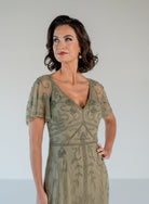 A woman modeling a sage green embroidered midi dress with sheer flutter sleeves and a V-neckline, set against a plain light gray studio background. / V neckline, soft flutter sleeve gown with godet skirt and all over beading.