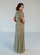 A woman modeling a floor-length sage green evening gown with sheer flutter sleeves and delicate floral embroidery, photographed against a plain light gray studio background. / V neckline, soft flutter sleeve gown with godet skirt and all over beading.
