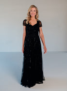 A woman modeling a floor-length black evening gown with sheer lace short sleeves and a V-neckline, standing against a plain light gray studio background. / Sweetheart neckline A-line gown with all over beading.