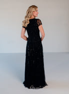 A woman modeling a floor-length black evening gown with lace short sleeves and a sequined tulle skirt, photographed from the back against a plain light gray studio backdrop. / Sweetheart neckline A-line gown with all over beading.