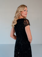 A blonde woman wearing a black sequined dress with sheer lace short sleeves, shown from a three-quarter back view against a light gray studio background. / Sweetheart neckline A-line gown with all over beading.