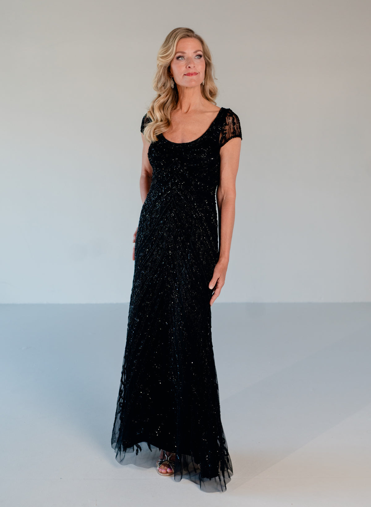 A woman modeling a floor-length black evening gown with sheer cap sleeves and a scoop neckline, standing against a plain light gray studio background. / Scoop neckline, slim gown with all over beading.