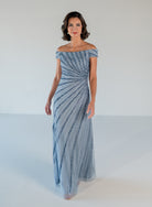 A woman modeling an off-the-shoulder floor-length gown with a textured, swirling striped pattern in light blue and white, set against a plain white studio background. / Off the shoulder slim gown with side rushing and linear beading.