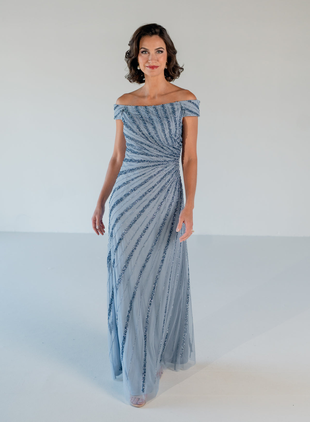A woman modeling an off-the-shoulder floor-length gown with a textured, swirling striped pattern in light blue and white, set against a plain white studio background. / Off the shoulder slim gown with side rushing and linear beading.