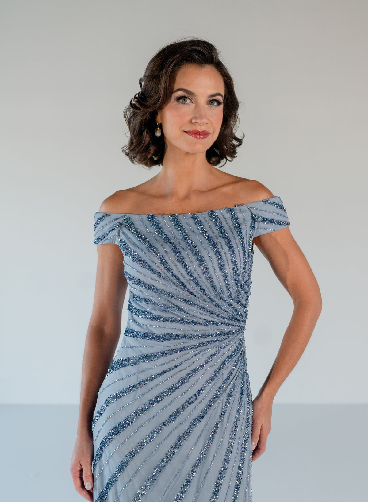 A woman modeling an off-the-shoulder evening gown with a textured, sparkly blue and silver striped pattern, set against a neutral studio background. / Off the shoulder slim gown with side rushing and linear beading.
