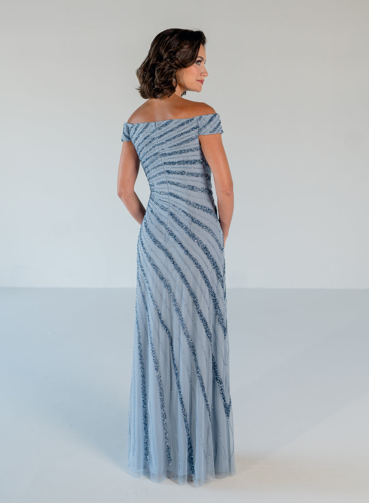 A woman modeling an off-the-shoulder floor-length gown with diagonal striped patterns in light blue and dark blue, shown from the back against a plain light gray studio background. / Off the shoulder slim gown with side rushing and linear beading.