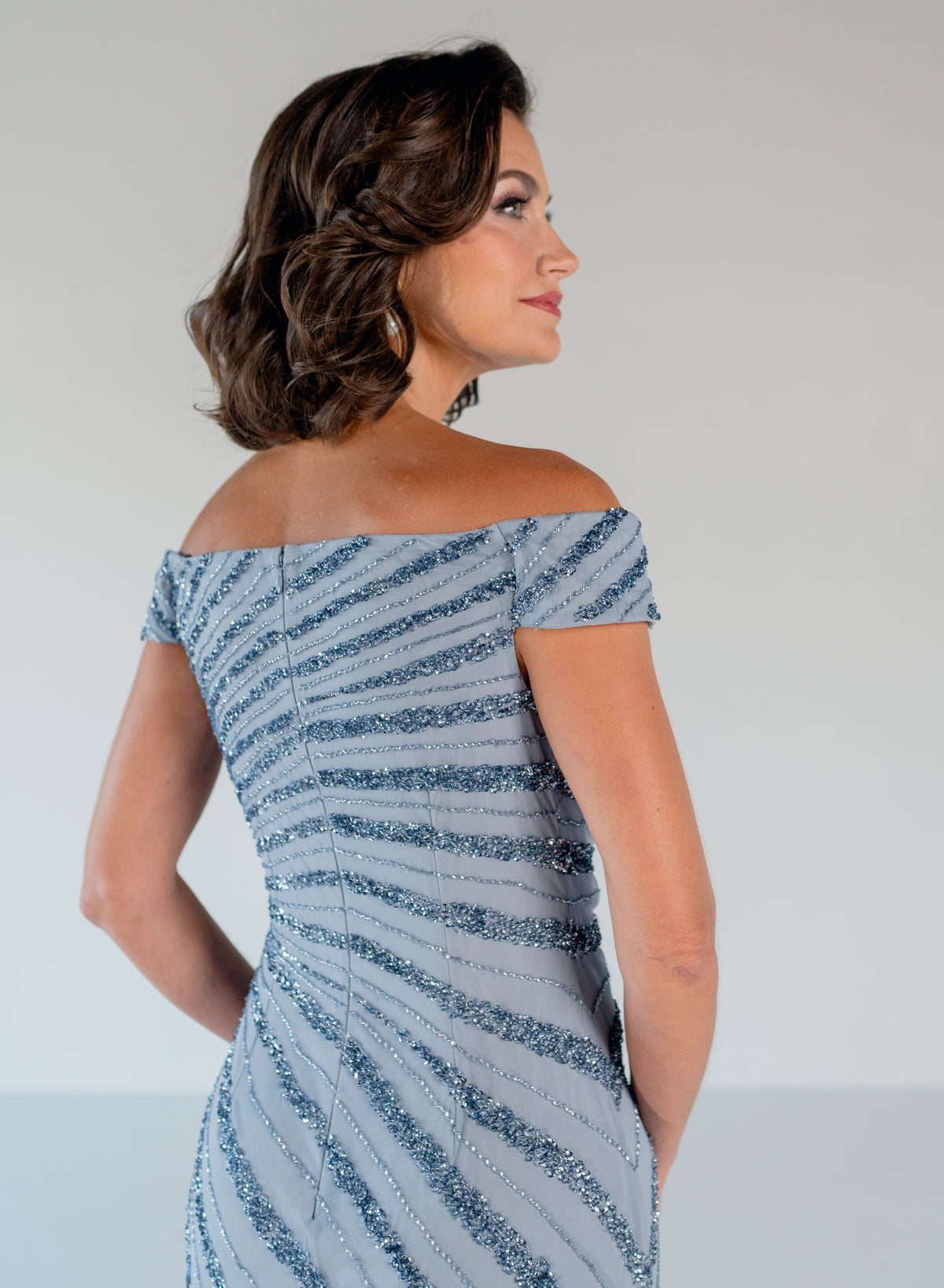 A woman shown from the back wearing an off-the-shoulder evening gown with diagonal silver glitter stripes on a light gray fabric, set against a plain light gray background. / Off the shoulder slim gown with side rushing and linear beading.