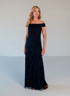 A woman modeling a floor-length off-the-shoulder evening gown in dark navy blue with shimmering vertical bead or sequin stripes, photographed against a plain light gray studio background. / Off the shoulder slim gown with side rushing and linear beading.