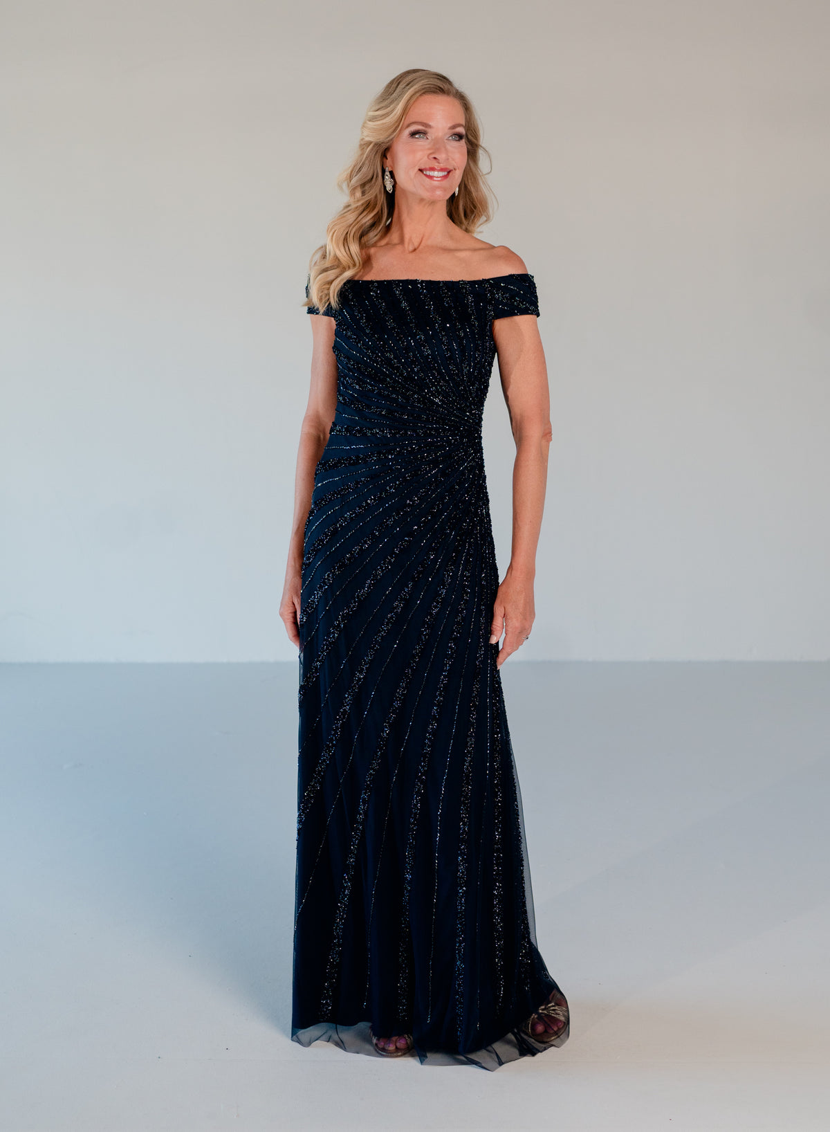 A woman modeling a floor-length off-the-shoulder evening gown in dark navy blue with shimmering vertical bead or sequin stripes, photographed against a plain light gray studio background. / Off the shoulder slim gown with side rushing and linear beading.