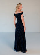 A woman modeling an off-the-shoulder, floor-length navy evening gown with subtle shimmering vertical stripes, photographed in a studio setting against a light gray background. / Off the shoulder slim gown with side rushing and linear beading.