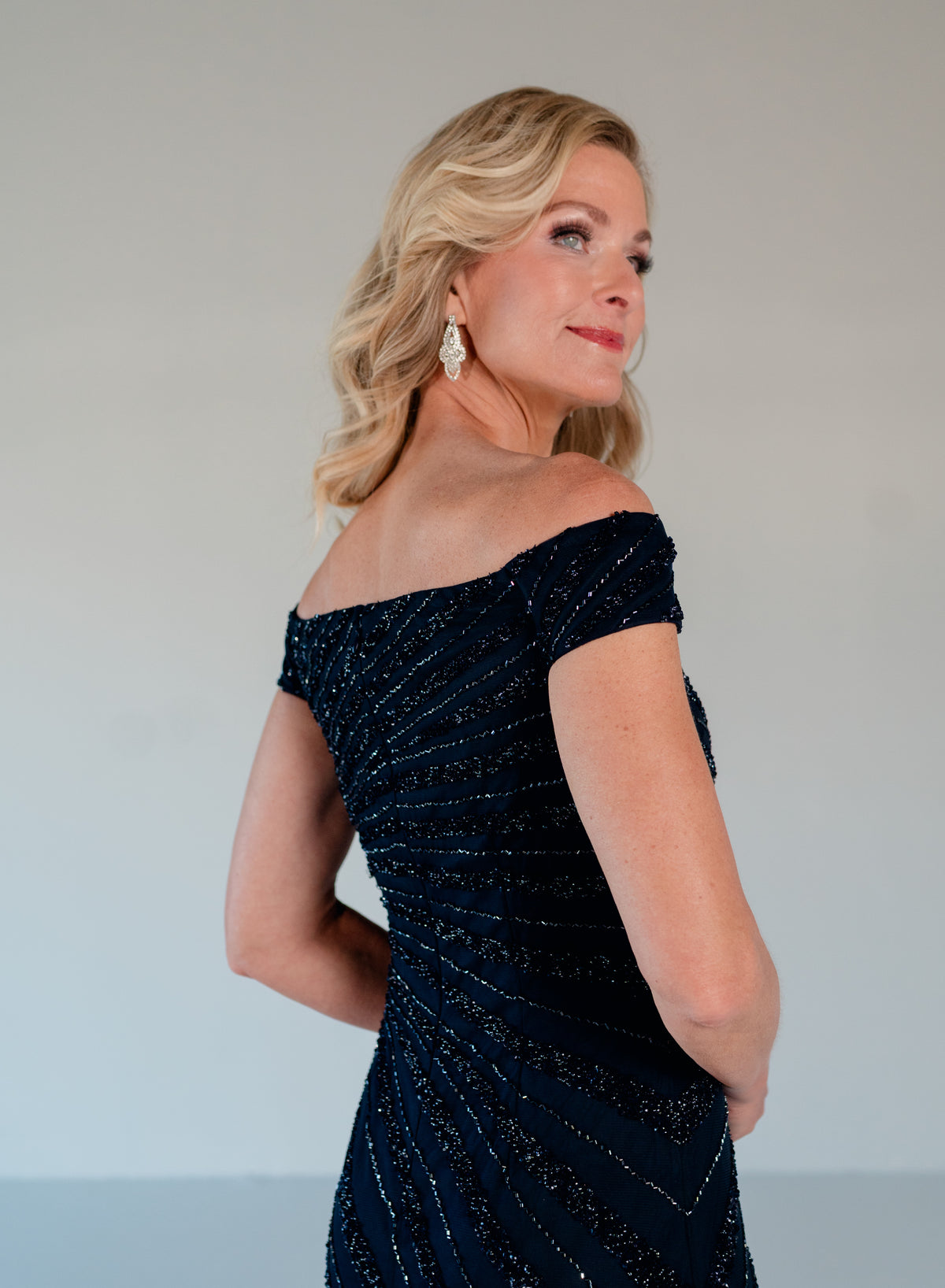 A blonde woman wearing an off-the-shoulder dark navy evening gown with shimmering sequined details, photographed against a plain light gray studio background. / Off the shoulder slim gown with side rushing and linear beading.