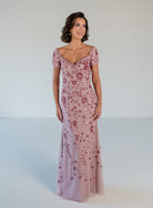 A woman modeling a floor-length dusty pink gown with short puff sleeves, a V-neckline, and an allover rose-gold sequin floral pattern against a light gray studio background. / Sweetheart neckline, slim gown with all over floral beading.