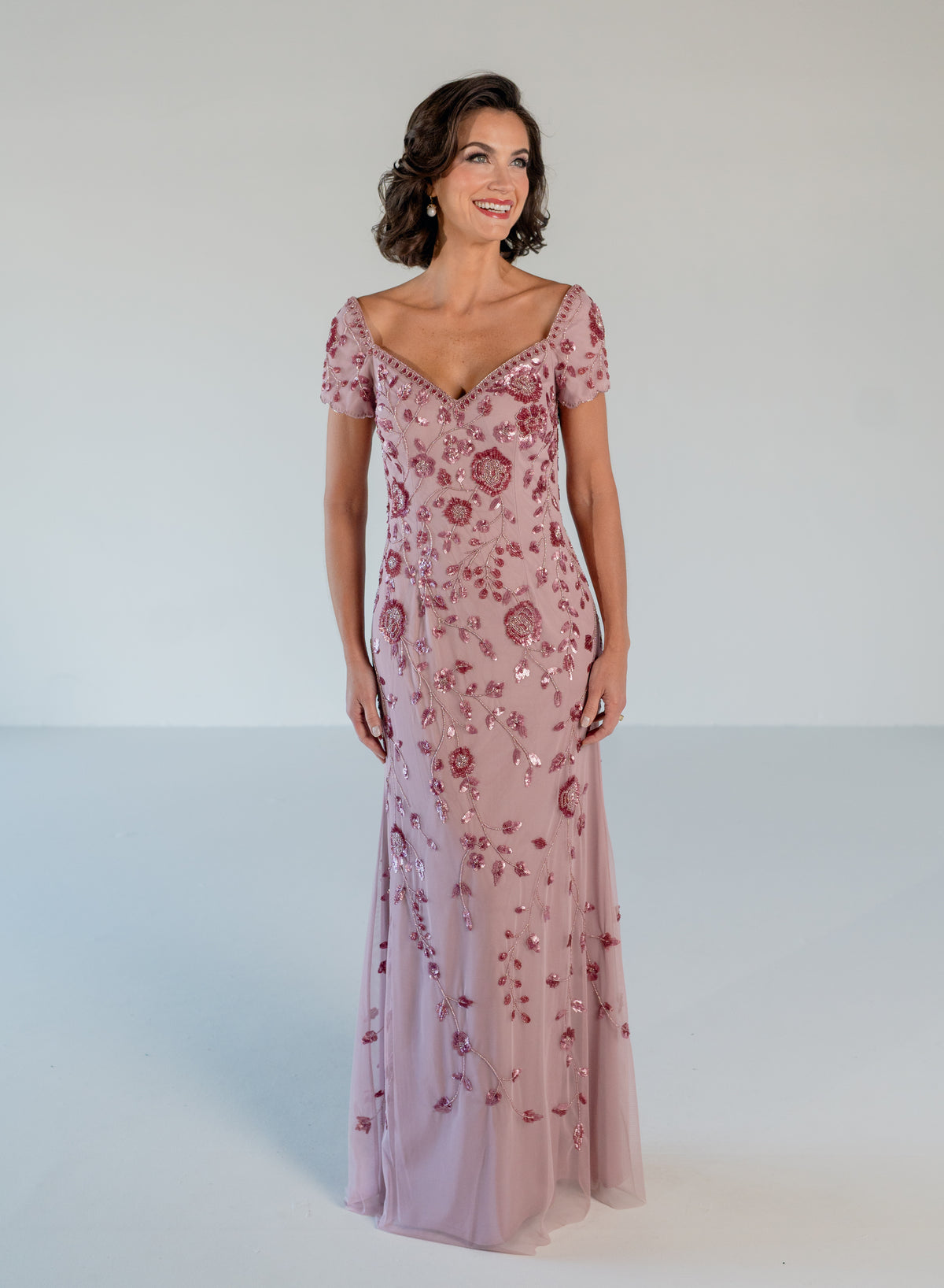 A woman modeling a floor-length dusty pink gown with short puff sleeves, a V-neckline, and an allover rose-gold sequin floral pattern against a light gray studio background. / Sweetheart neckline, slim gown with all over floral beading.