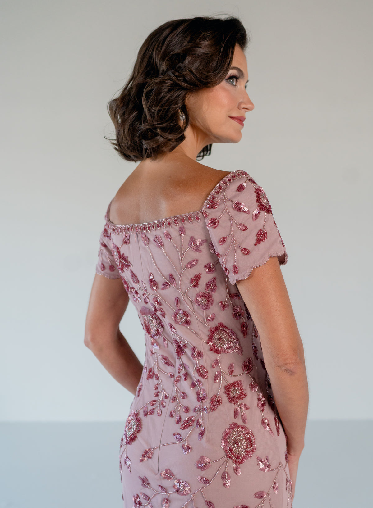 A woman shown from the back wearing a dusty pink off-the-shoulder evening gown decorated with rose pink floral sequins and beadwork, set against a plain light gray studio background. / Sweetheart neckline, slim gown with all over floral beading.