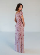 A woman modeling a floor-length dusty pink gown with short puff sleeves and an allover rose floral sequin and bead embellishment, photographed against a seamless light gray studio background. / Sweetheart neckline, slim gown with all over floral beading.