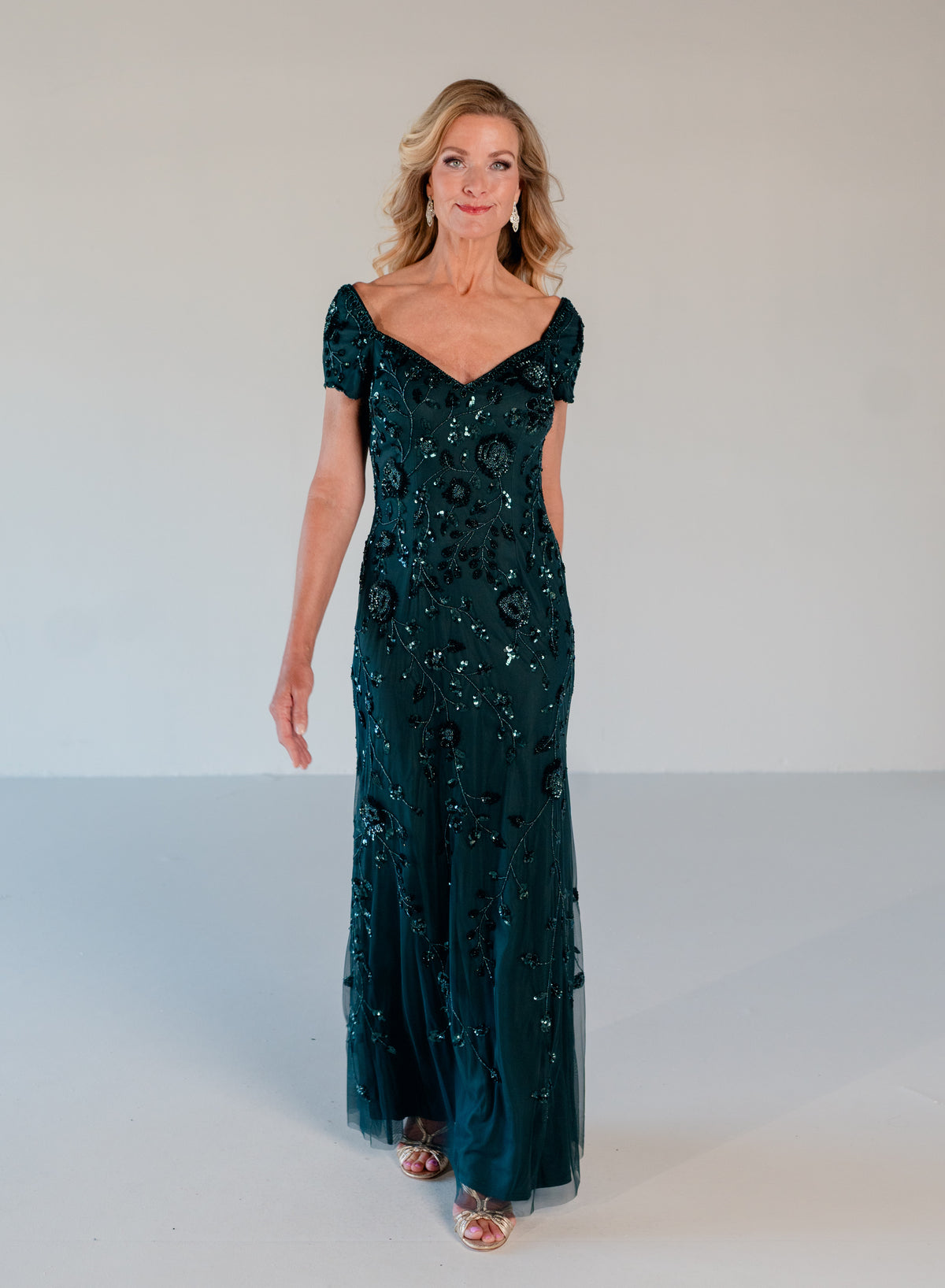 A woman modeling a floor-length dark teal green evening gown with short puff sleeves, a V-neckline, and dense sequined floral patterns, set against a plain light gray studio background. / Sweetheart neckline, slim gown with all over floral beading.