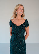 A woman modeling a dark teal green off-the-shoulder evening gown with intricate floral sequin and bead embroidery, set against a neutral light gray studio background. / Sweetheart neckline, slim gown with all over floral beading.