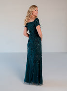 A woman modeling a floor-length off-the-shoulder gown in deep teal with intricate black beaded floral patterns, photographed against a neutral light gray studio background. / Sweetheart neckline, slim gown with all over floral beading.