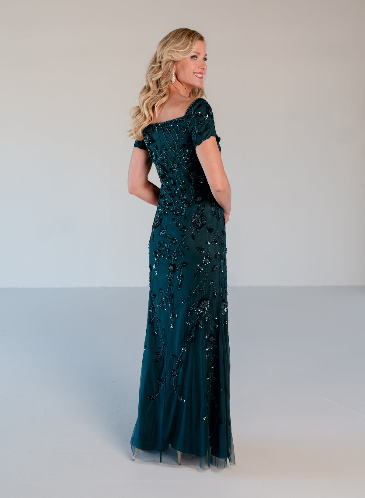 A woman modeling a floor-length off-the-shoulder gown in deep teal with intricate black beaded floral patterns, photographed against a neutral light gray studio background. / Sweetheart neckline, slim gown with all over floral beading.