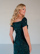 A woman modeling an off-the-shoulder dark teal green evening gown with intricate sequin and bead floral embroidery, shown from a three-quarter back view against a neutral studio background. / Sweetheart neckline, slim gown with all over floral beading.