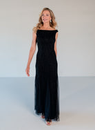 A woman modeling a sleeveless, off-the-shoulder black evening gown with beaded detailing and a flowing tulle skirt, standing against a plain light gray studio background. / Off the shoulder slim gown with soft godets and all-over linear beading.