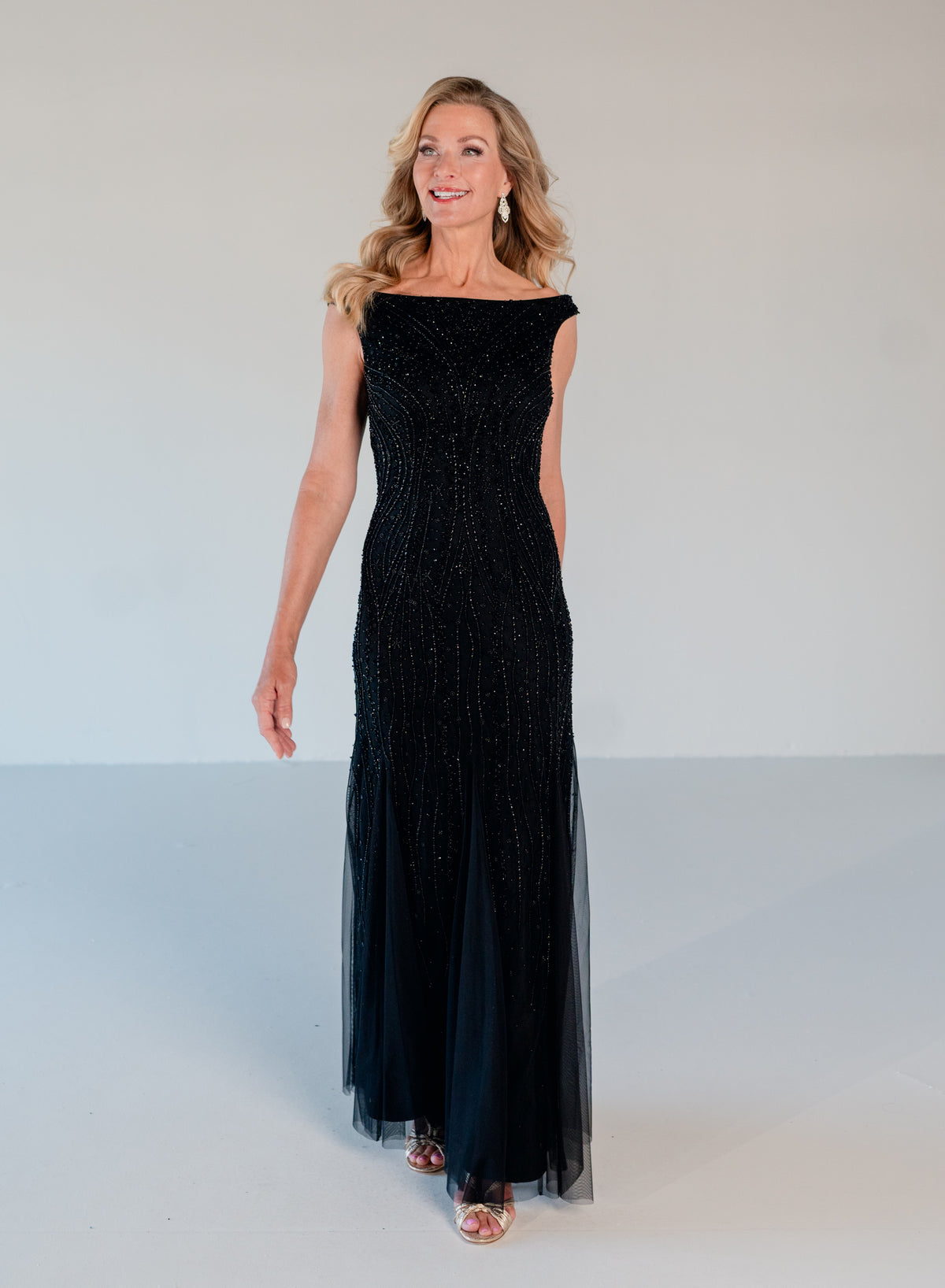A woman modeling a sleeveless, off-the-shoulder black evening gown with beaded detailing and a flowing tulle skirt, standing against a plain light gray studio background. / Off the shoulder slim gown with soft godets and all-over linear beading.