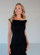 A woman wearing a sleeveless black beaded off-the-shoulder evening gown, standing against a neutral light gray studio background. / Off the shoulder slim gown with soft godets and all-over linear beading.