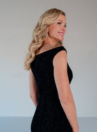 A blonde woman wearing a sleeveless black beaded dress, shown from a side-back angle against a plain light gray studio background. / Off the shoulder slim gown with soft godets and all-over linear beading.