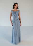 A woman modeling a floor-length light blue gown with a fitted bodice, off-the-shoulder neckline, and cascading beaded embellishments, set against a plain white studio background. / Off the shoulder slim gown with soft godets and all-over linear beading.