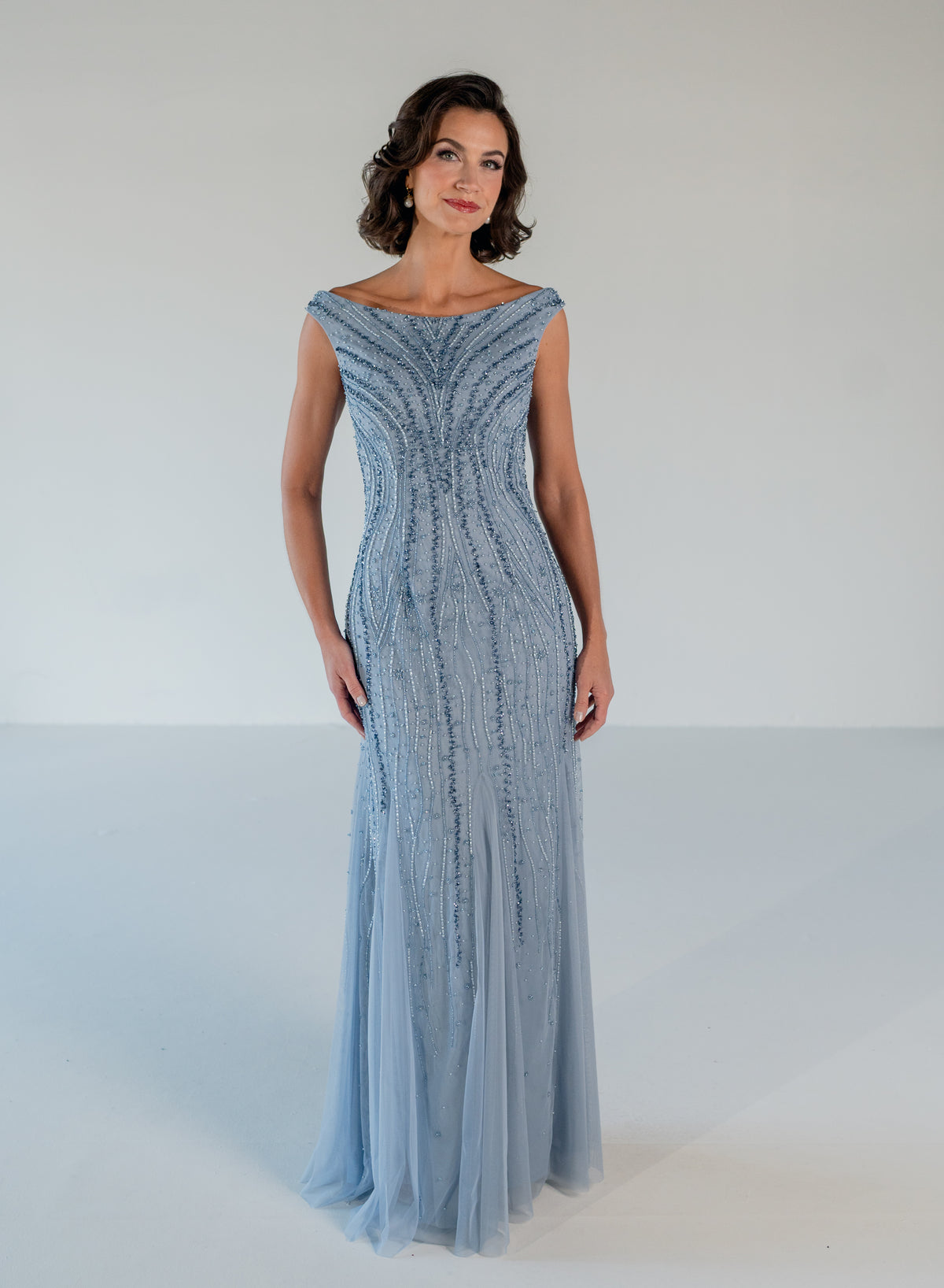 A woman modeling a floor-length light blue gown with a fitted bodice, off-the-shoulder neckline, and cascading beaded embellishments, set against a plain white studio background. / Off the shoulder slim gown with soft godets and all-over linear beading.