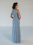 A woman modeling a floor-length light blue evening gown with vertical beaded detailing and a sheer tulle skirt, photographed against a plain light gray studio background. / Off the shoulder slim gown with soft godets and all-over linear beading.