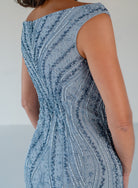 Close-up back view of a woman wearing a fitted sleeveless dress with a dusty blue base and dense vertical beaded and sequined stripes in matching blue and silver tones. / Off the shoulder slim gown with soft godets and all-over linear beading.