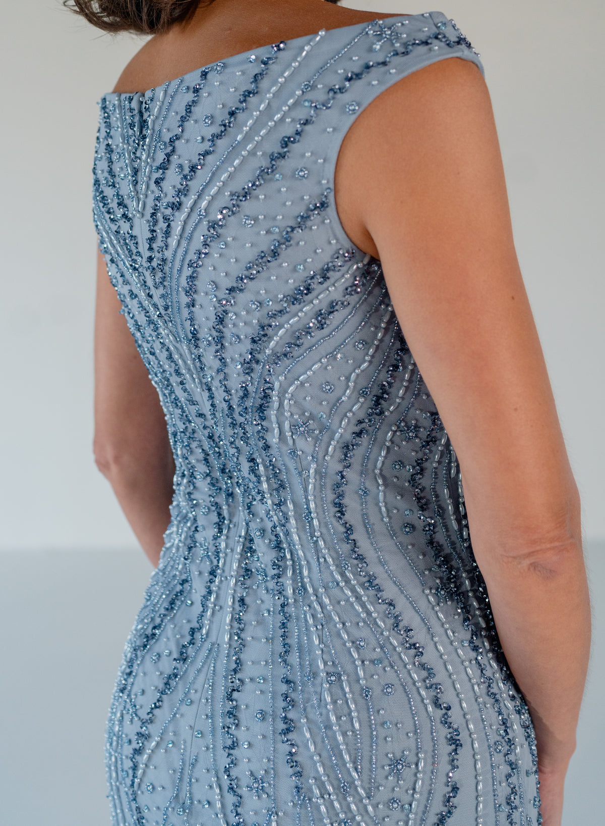 Close-up back view of a woman wearing a fitted sleeveless dress with a dusty blue base and dense vertical beaded and sequined stripes in matching blue and silver tones. / Off the shoulder slim gown with soft godets and all-over linear beading.