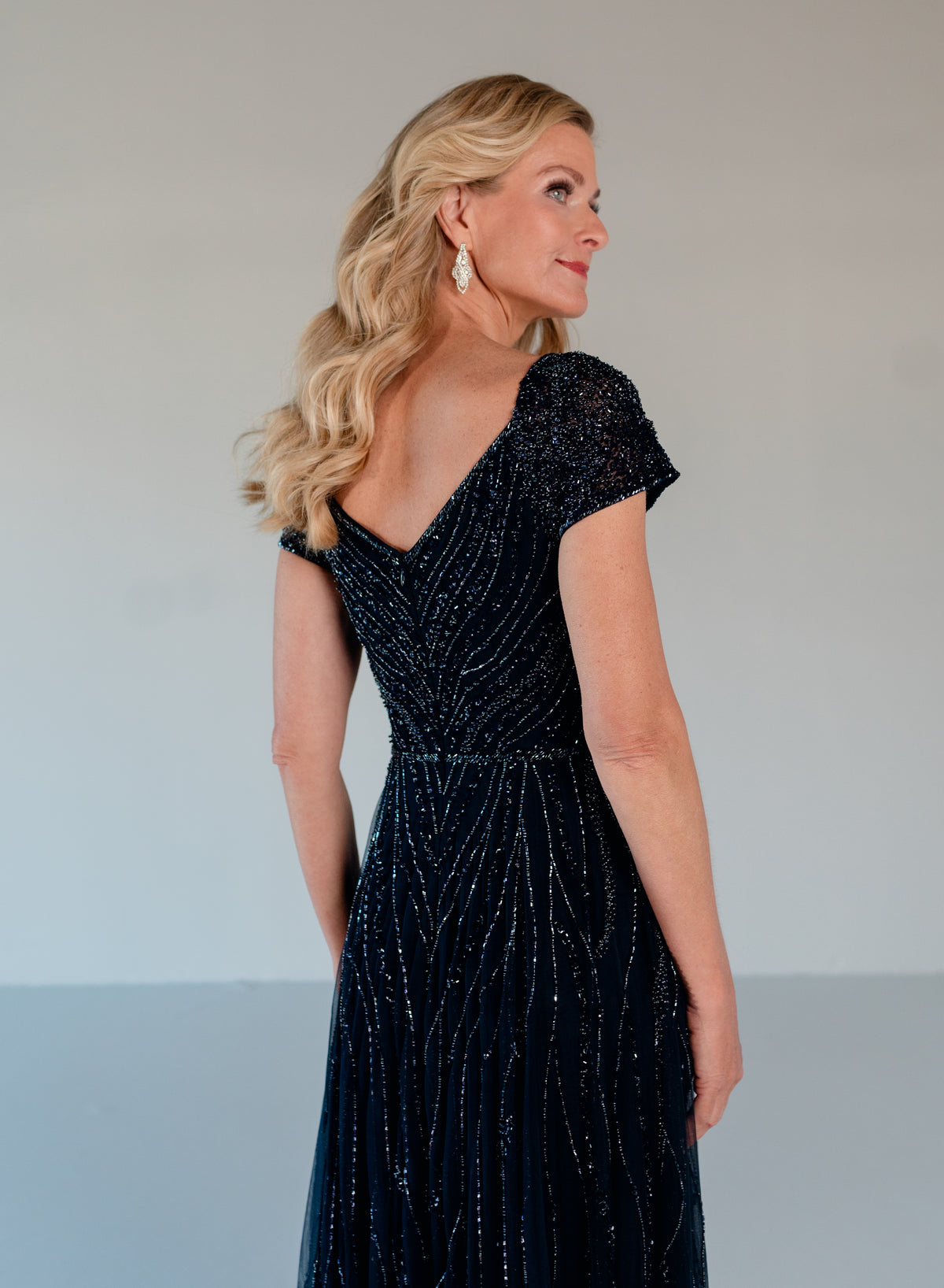 A blonde woman wearing a dark navy blue evening gown with sparkling vertical bead and sequin details, shown from a three-quarter back view against a neutral studio background. / Sweetheart neckline-line gown with all over trailing beading.