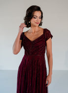 A woman wearing a floor-length burgundy evening gown with sparkling beadwork and cap sleeves, photographed against a plain light gray studio background. / Sweetheart neckline-line gown with all over trailing beading.