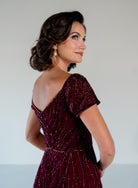 A woman wearing a deep burgundy off-the-shoulder evening gown with intricate beadwork and sequins, shown from a three-quarter back view against a light gray studio background. / Sweetheart neckline-line gown with all over trailing beading.