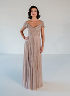 A woman modeling a floor-length evening gown in a light blush beige color. The dress features a lace bodice with short cap sleeves, a V-neckline, and a flowing tulle skirt, set against a plain white studio background. / Sweetheart neckline-line gown with all over trailing beading.