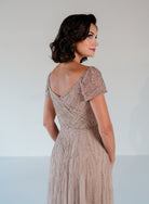 A woman wearing a champagne beige evening gown with intricate beadwork and short puff sleeves, shown from a three-quarter back view against a neutral studio background. / Sweetheart neckline-line gown with all over trailing beading.