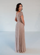 A woman modeling a floor-length, off-the-shoulder evening gown in a dusty blush pink or taupe-beige tone. The dress features intricate lace and bead embellishments on the bodice and sleeves, with a flowing tulle skirt, shown against a plain white studio background. / Sweetheart neckline-line gown with all over trailing beading.