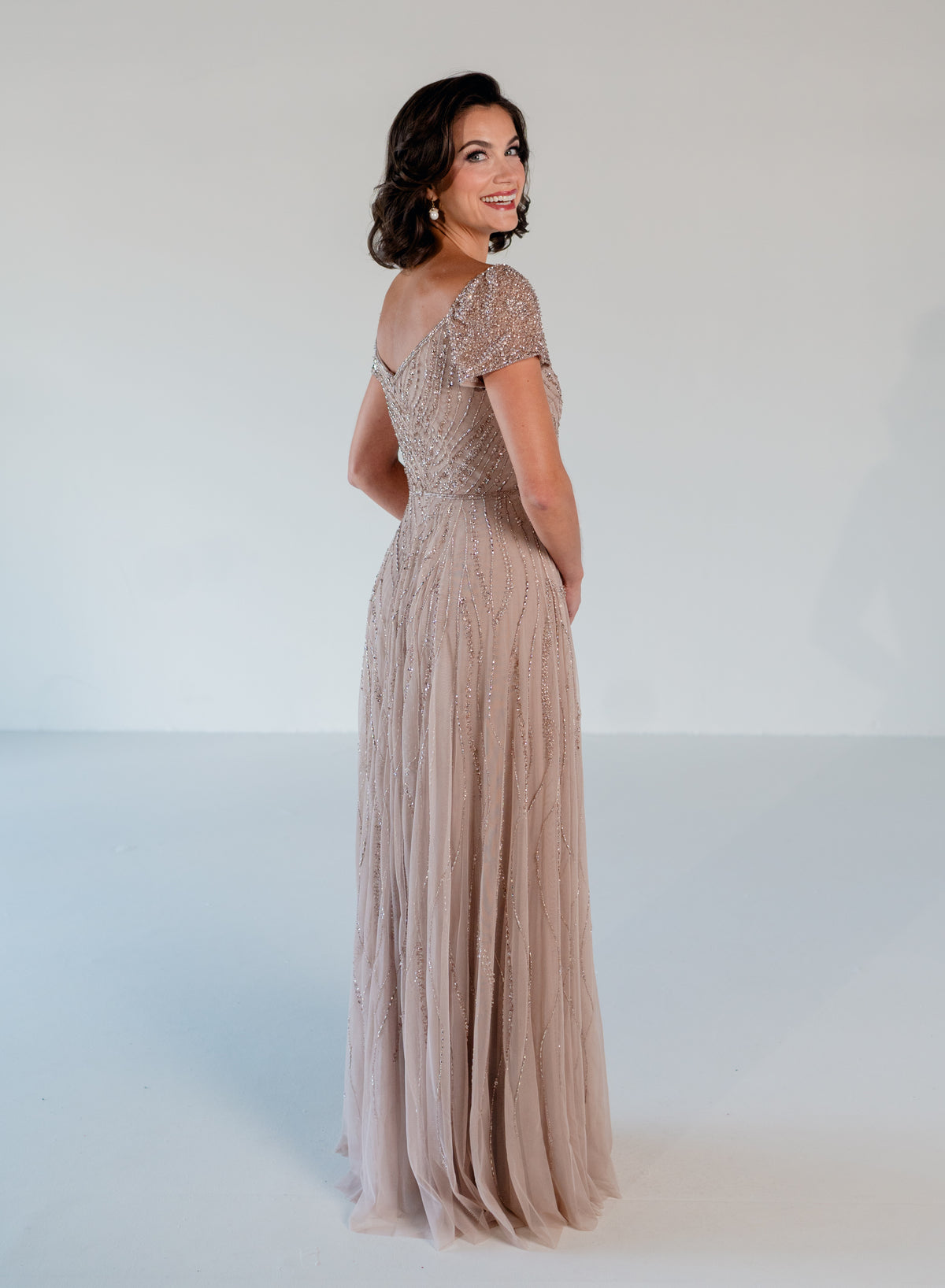 A woman modeling a floor-length, off-the-shoulder evening gown in a dusty blush pink or taupe-beige tone. The dress features intricate lace and bead embellishments on the bodice and sleeves, with a flowing tulle skirt, shown against a plain white studio background. / Sweetheart neckline-line gown with all over trailing beading.