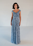 A woman modeling a floor-length light blue evening gown with silver beadwork and sequins, photographed against a plain white studio background. / Off the shoulder A-line gown with all over floral and trailing beading.