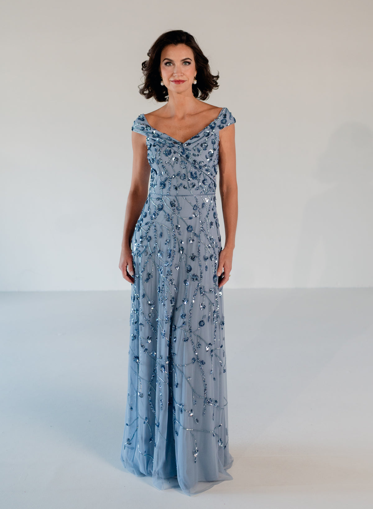 A woman modeling a floor-length light blue evening gown with silver beadwork and sequins, photographed against a plain white studio background. / Off the shoulder A-line gown with all over floral and trailing beading.